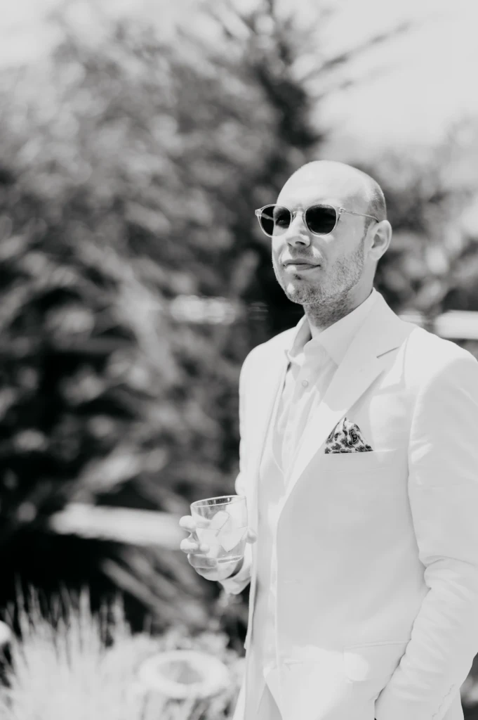 Groom Stavros standing in a white suit with a drink, black and white