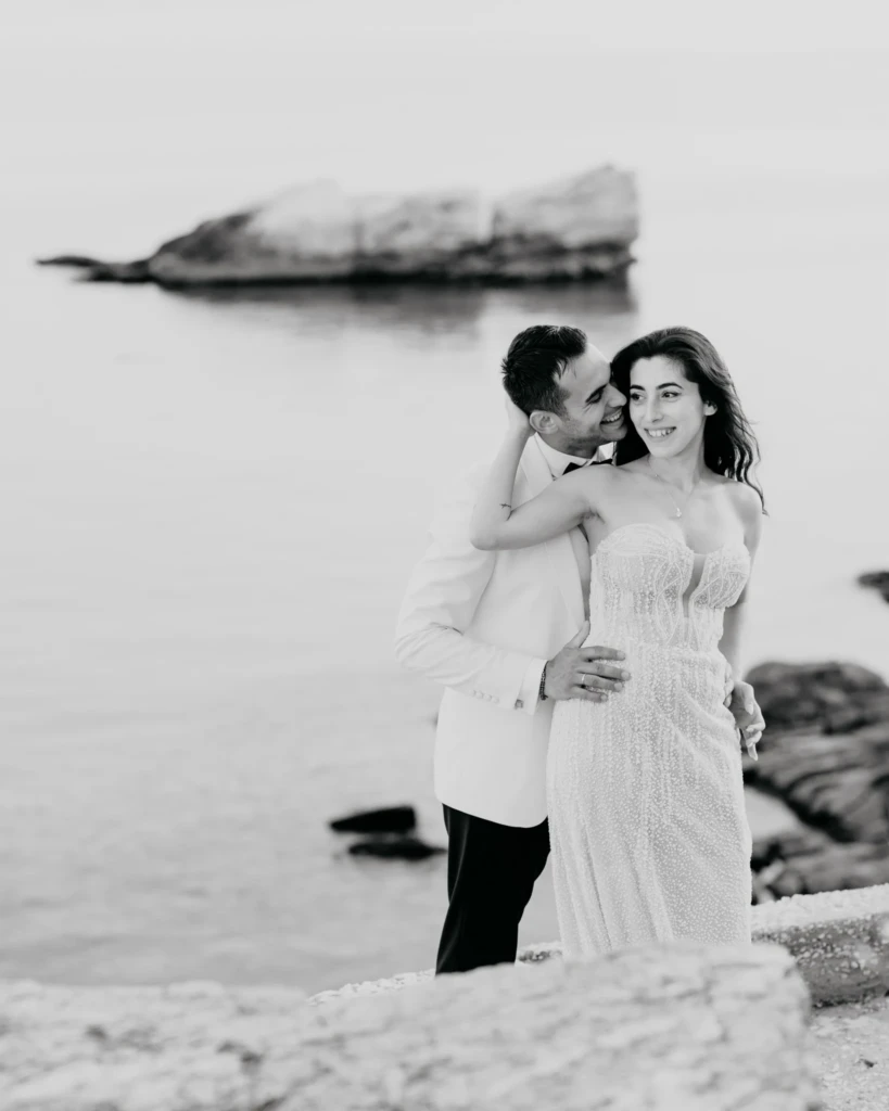 Anastasia and Aggelos hand in hand on the rocks by the sea after their wedding