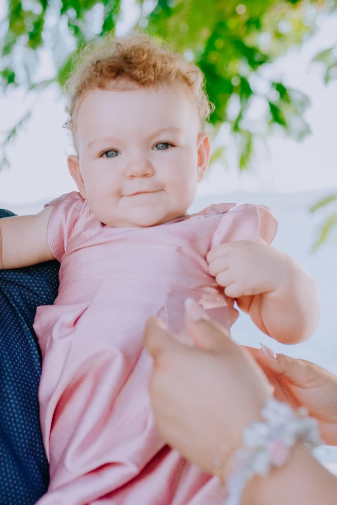 Close portrait of a curly-haired baby in a pink dress, held under green leaves at a christening