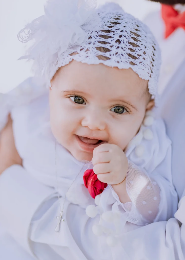 Smiling baby in a white lace christening bonnet and gown, holding a small silver cross