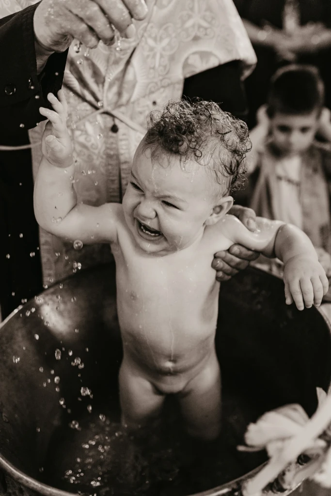 Black and white photo of a baby standing in the baptismal font as the Orthodox priest pours blessed water, droplets in the air