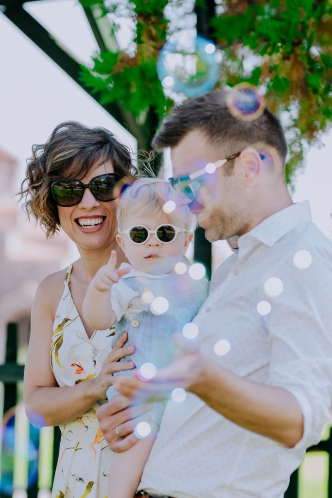 Parents laughing with their toddler in white sunglasses among floating soap bubbles at a christening celebration