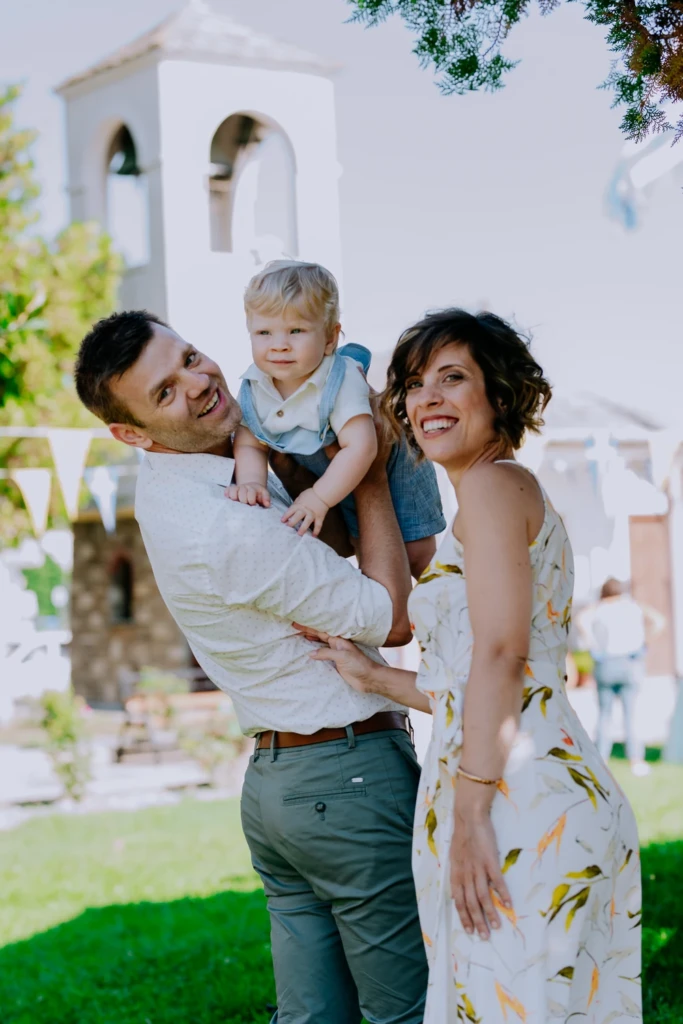Mother and father laughing as they lift their baby boy in front of a white church bell tower after his christening