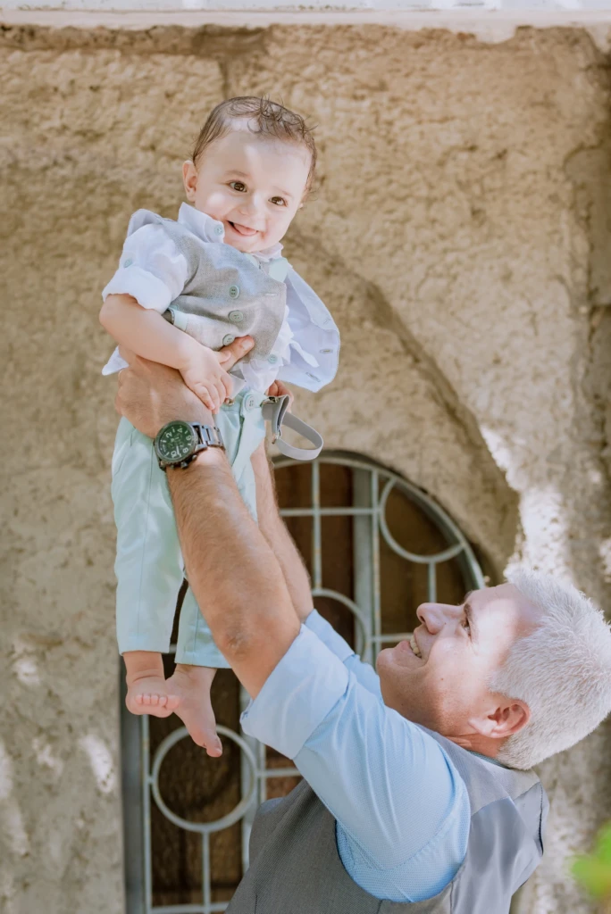 Silver-haired grandfather lifting a smiling baby boy high in the air after his baptism, the child's hair still wet