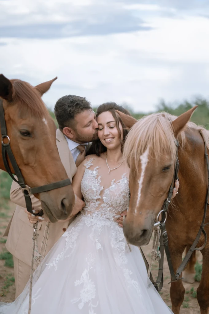 Vasilis kissing Elena's cheek as she smiles, standing between two horses in the countryside
