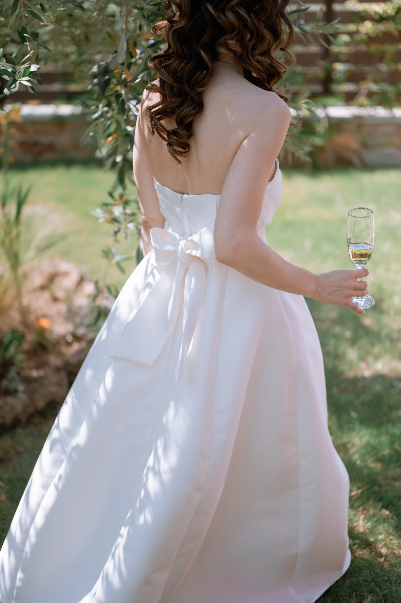 Bride Eva from behind in her gown holding a champagne glass among the greenery