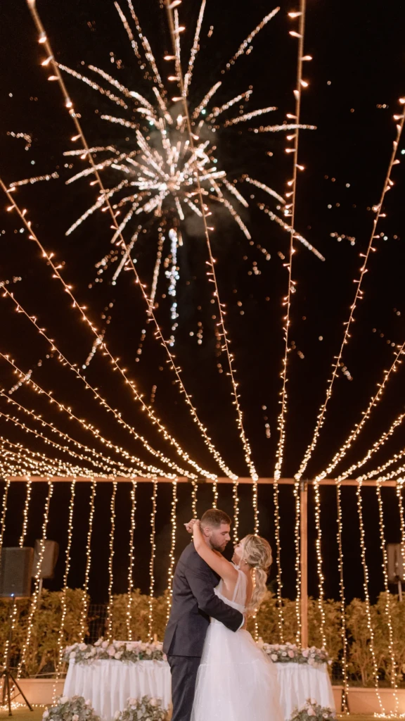 Fenia and Dimitris embracing under fireworks and fairy lights at night