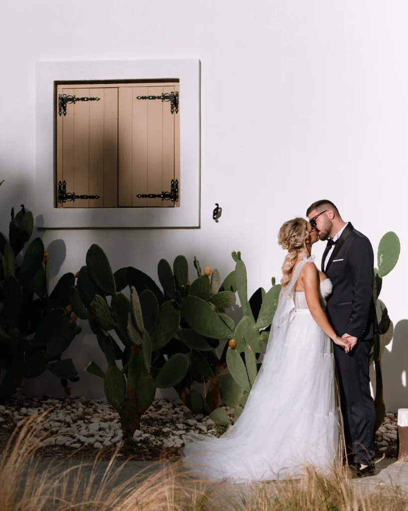 Fenia and Dimitris kissing beside a whitewashed wall and cacti