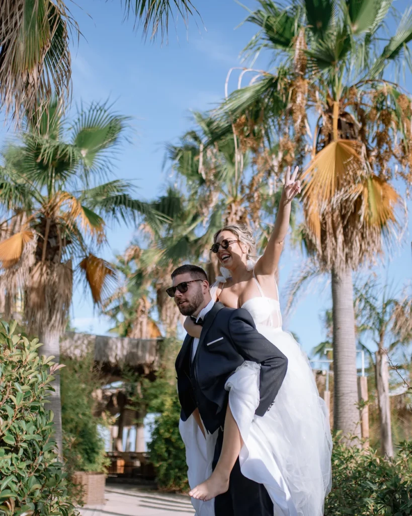 Dimitris giving Fenia a piggyback ride under palm trees, both laughing