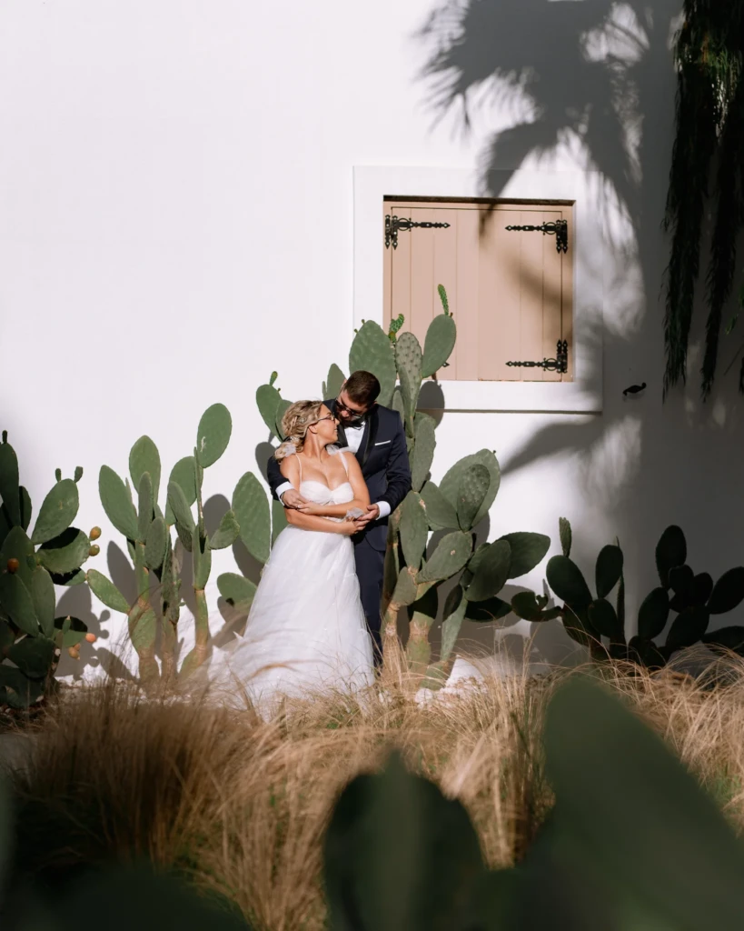 Fenia and Dimitris walking past cacti and a whitewashed wall in their wedding attire