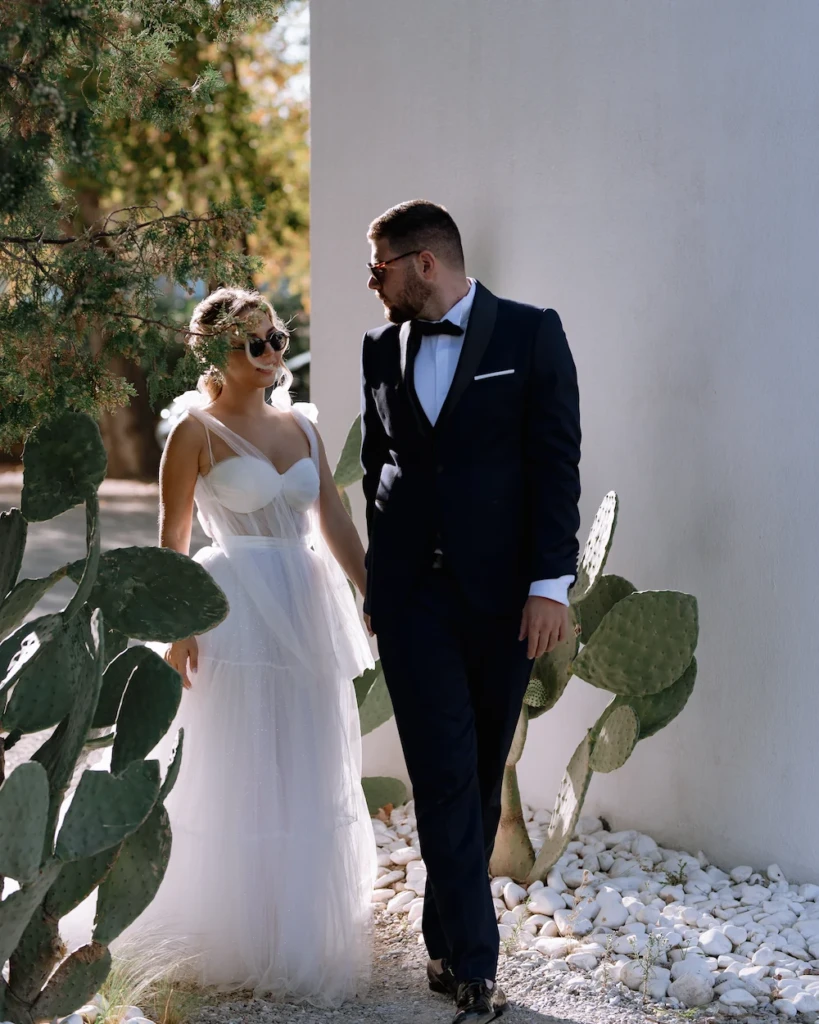 Fenia and Dimitris walking along a whitewashed wall framed by cacti and cypress trees