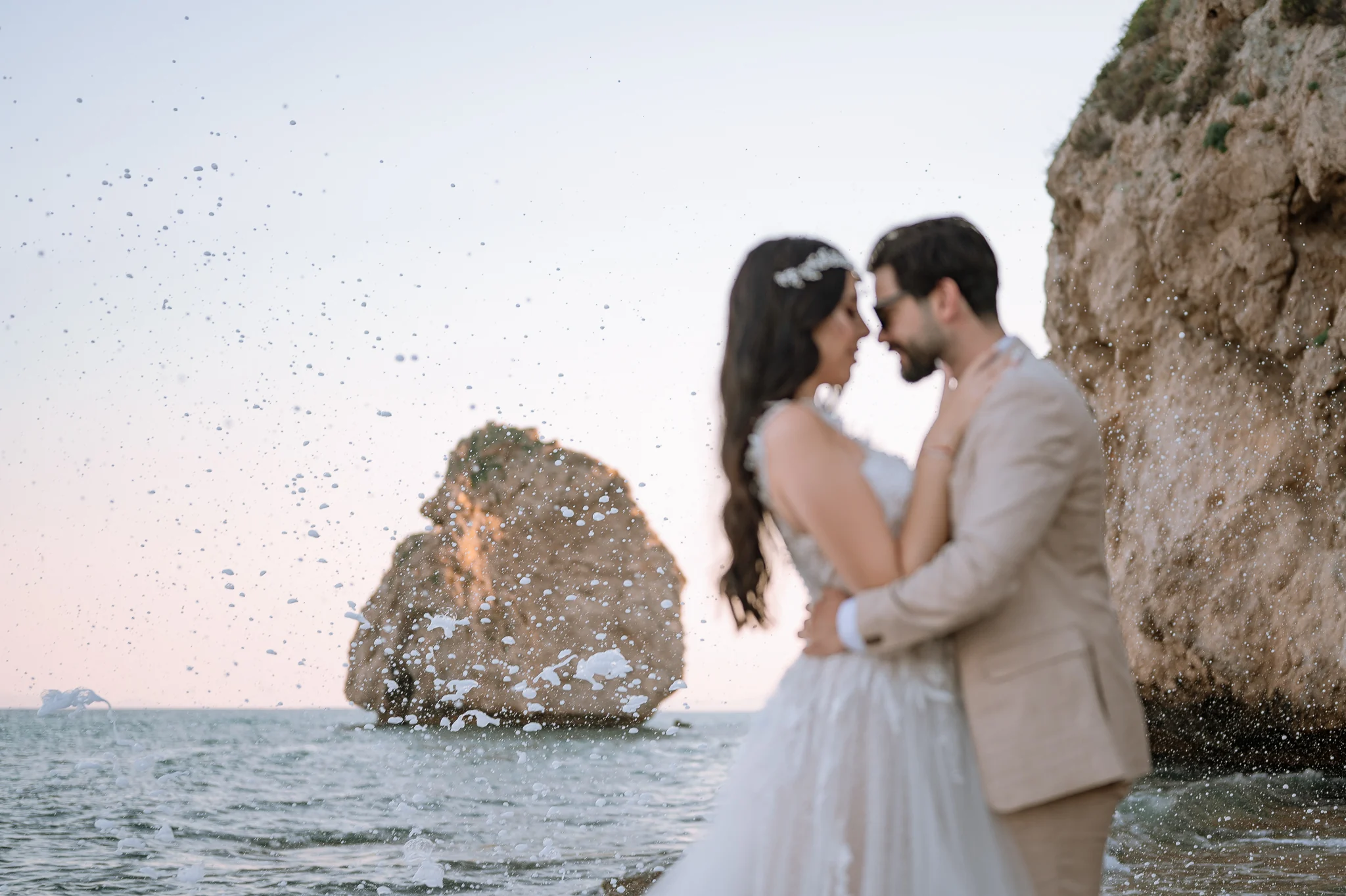 Bride and groom embracing, forehead to forehead, on the beach at sunset, with waves crashing against the rocks — wedding photoshoot in Kavala.