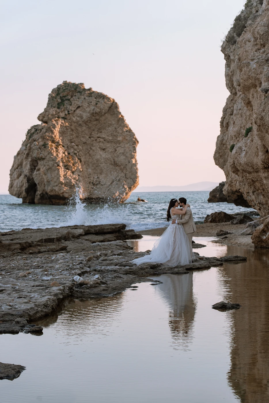 Νύφη και γαμπρός αγκαλιασμένοι σε βραχώδη παραλία την ώρα του δειλινού, με την αντανάκλασή τους στο νερό — γαμήλια φωτογράφιση στην ακτή της Καβάλας