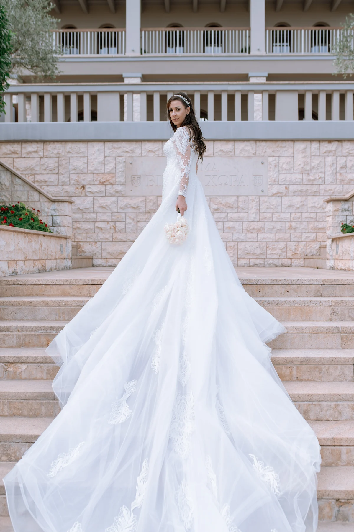 Bride Loukiani glancing back on the grand staircase at Ktima Biblia Chora, her tulle train spread out behind her