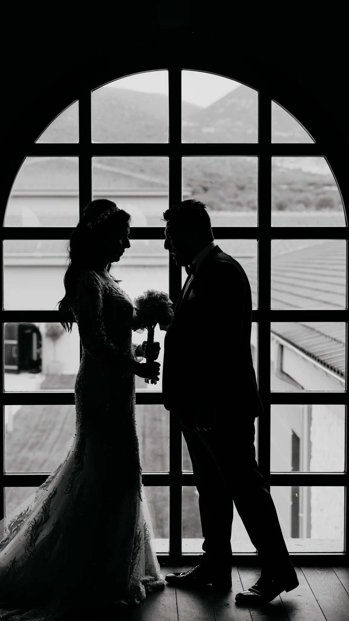 Loukiani and Giorgos silhouetted at a tall arched window over the Biblia Chora vineyards, bride holding her bouquet