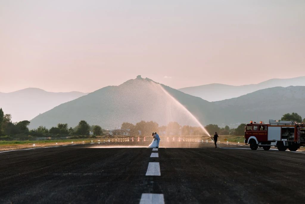 Newlywed couple embracing on an airport runway with a fire truck and water in the golden light of dusk, mountains in the background — wedding photoshoot by Moments of Art, Kavala.